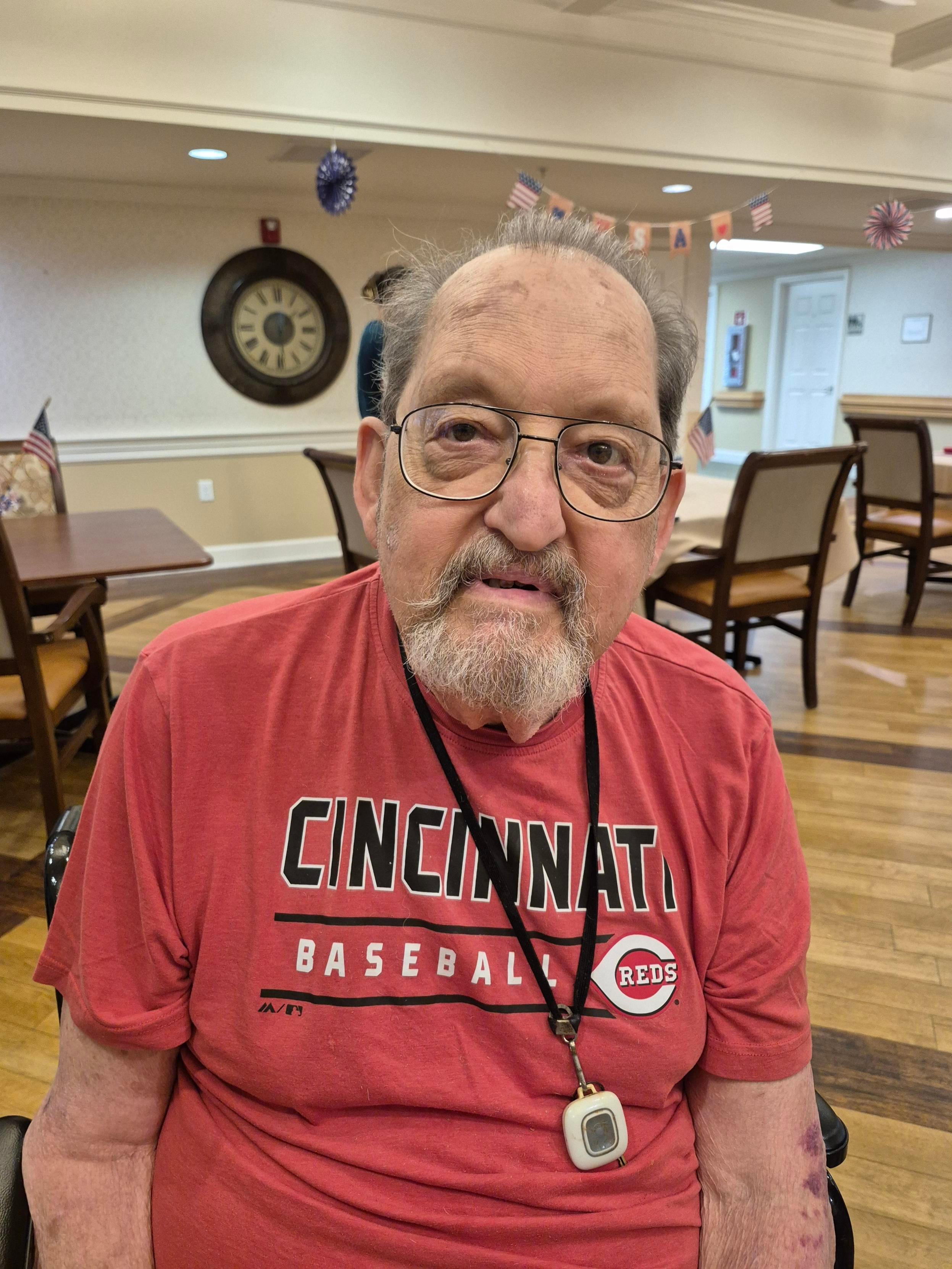 An older man wearing a Cincinnati Reds baseball shirt sits in a community gathering space, looking toward the camera. Seated in a wheelchair, he is surrounded by tables, chairs, and patriotic decorations, creating a warm and welcoming community atmosphere.