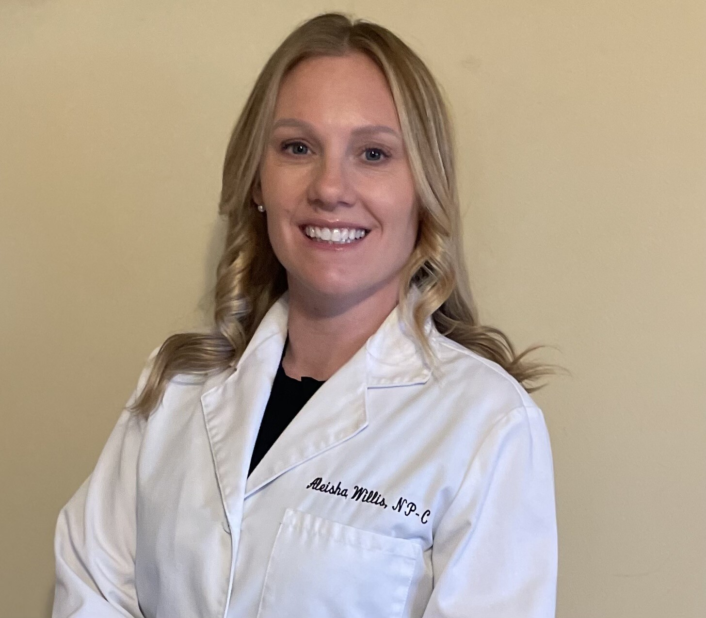 Professional portrait of Aleisha, NP-C, smiling at the camera. She has shoulder-length blonde hair and wears a white medical coat embroidered with her name and credentials over a black top, standing against a neutral beige background.