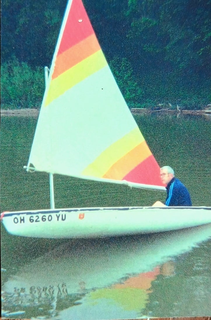 Older man sails a small white sailboat across a calm lake. The boat features a colorful striped sail, with a wooded shoreline visible in the background.