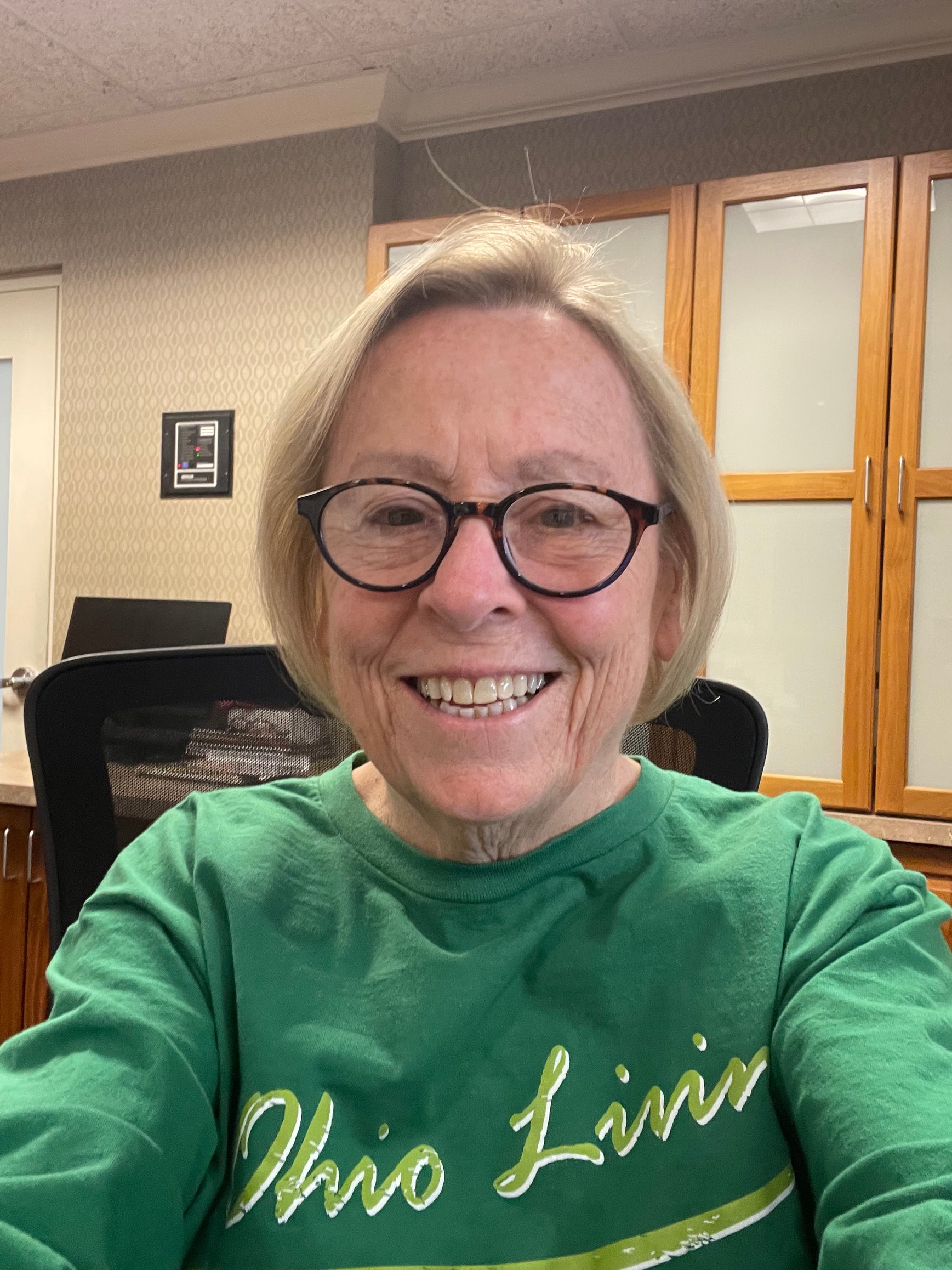 A smiling older woman with short blonde hair and round glasses sits indoors, facing the camera. She wears a green Ohio Living shirt. Behind her are wood cabinets, a chair, and a neutral office setting with soft lighting.