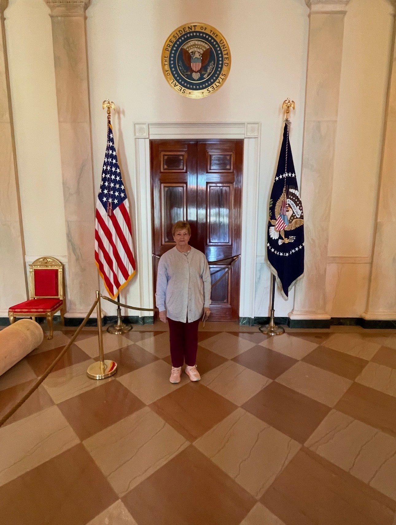 An older woman stands beneath the Seal of the President of the United States, between the American flag and presidential flag, in an elegant ceremonial setting with marble columns and a large wooden doorway.