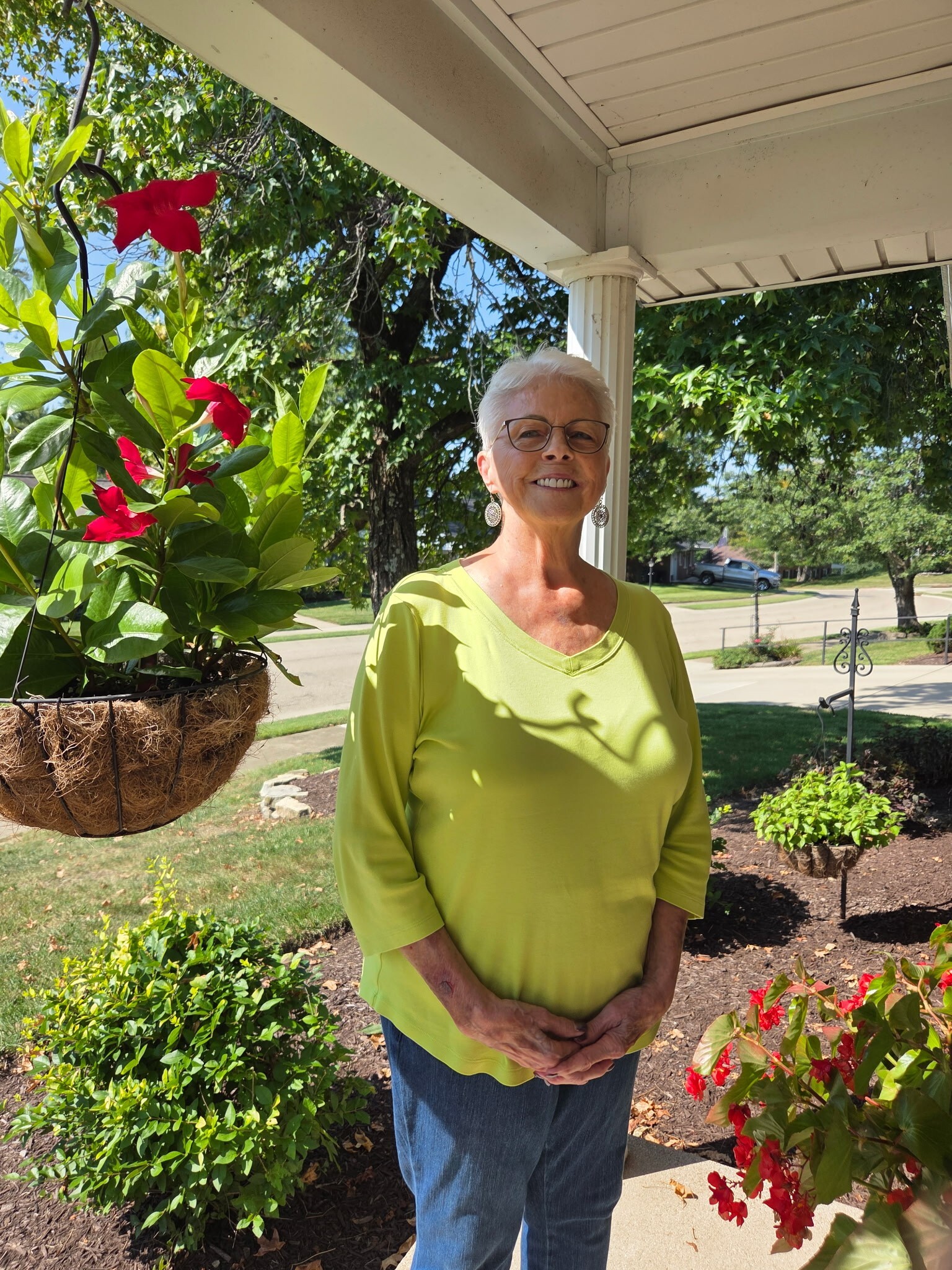 Smiling older woman with short white hair and glasses stands on a sunny porch surrounded by green plants and bright red flowers.