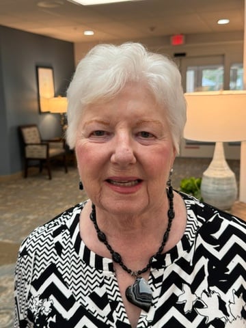 An older woman with short white hair smiles warmly indoors, wearing a black-and-white patterned top and a black necklace. Soft lighting and a welcoming community setting create a friendly portrait that reflects warmth, wisdom, and a caring personality.