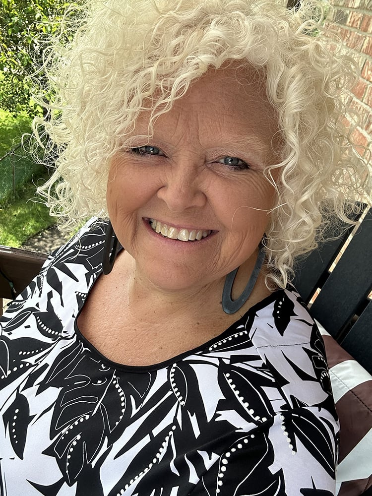 A smiling older woman with curly white hair and large black earrings poses outdoors, wearing a black-and-white floral patterned top.