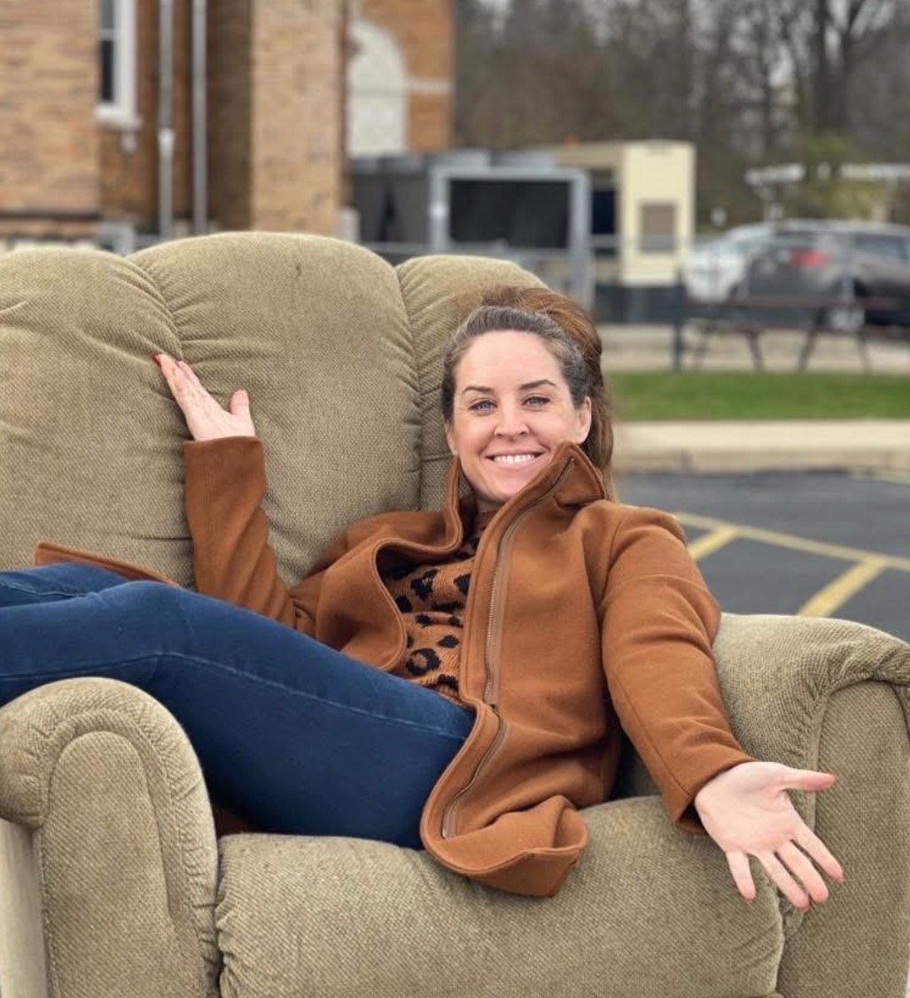 Woman smiling while relaxing on a large recliner placed outdoors in a parking lot. She is wearing a camel-colored jacket, patterned top, and dark jeans, playfully posing with open arms to showcase the oversized chair in a fun and inviting way.