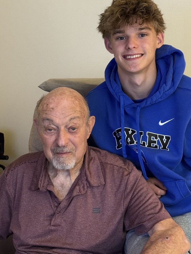 Older man with a white goatee wearing a maroon striped polo shirt seated beside a smiling teenage boy in a blue hoodie. The pair pose together indoors, capturing a warm, multigenerational family moment.