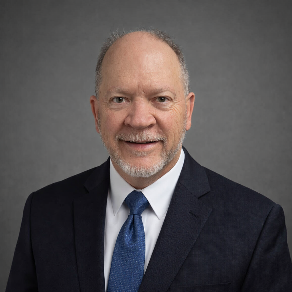 Professional headshot of a smiling man with a closely trimmed gray beard and mustache, wearing a navy suit, white shirt, and blue tie. He is posed against a neutral gray background, creating a polished, approachable portrait suitable for business or leadership profiles.