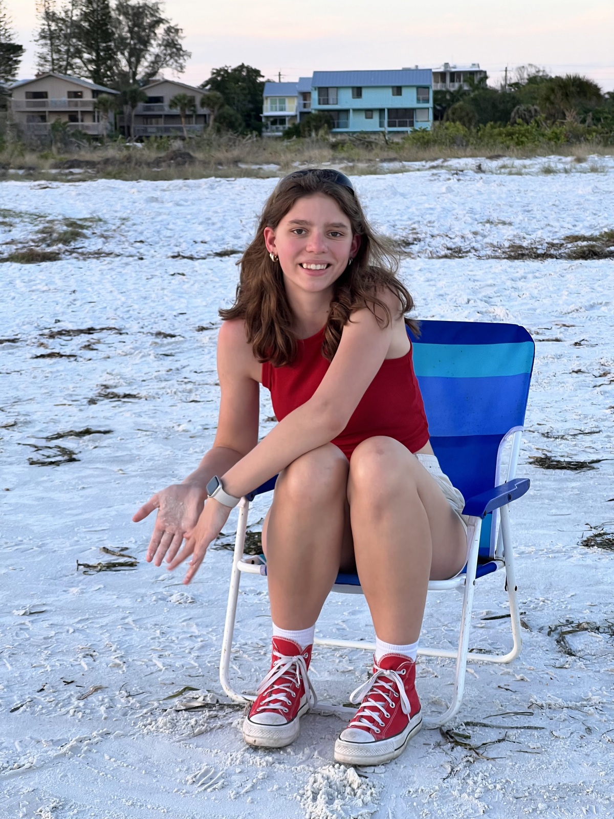 A young person sits on a low blue beach chair on a sandy shoreline at sunset, facing the camera and smiling. They are wearing a red sleeveless top, light-colored shorts, white socks, and red canvas sneakers. A smartwatch is visible on their left wrist, and they have small earrings. Their left hand is extended slightly forward, holding a bit of sand. The beach is scattered with small patches of seaweed, and behind them are several coastal houses, trees, and low vegetation under a soft evening sky.
