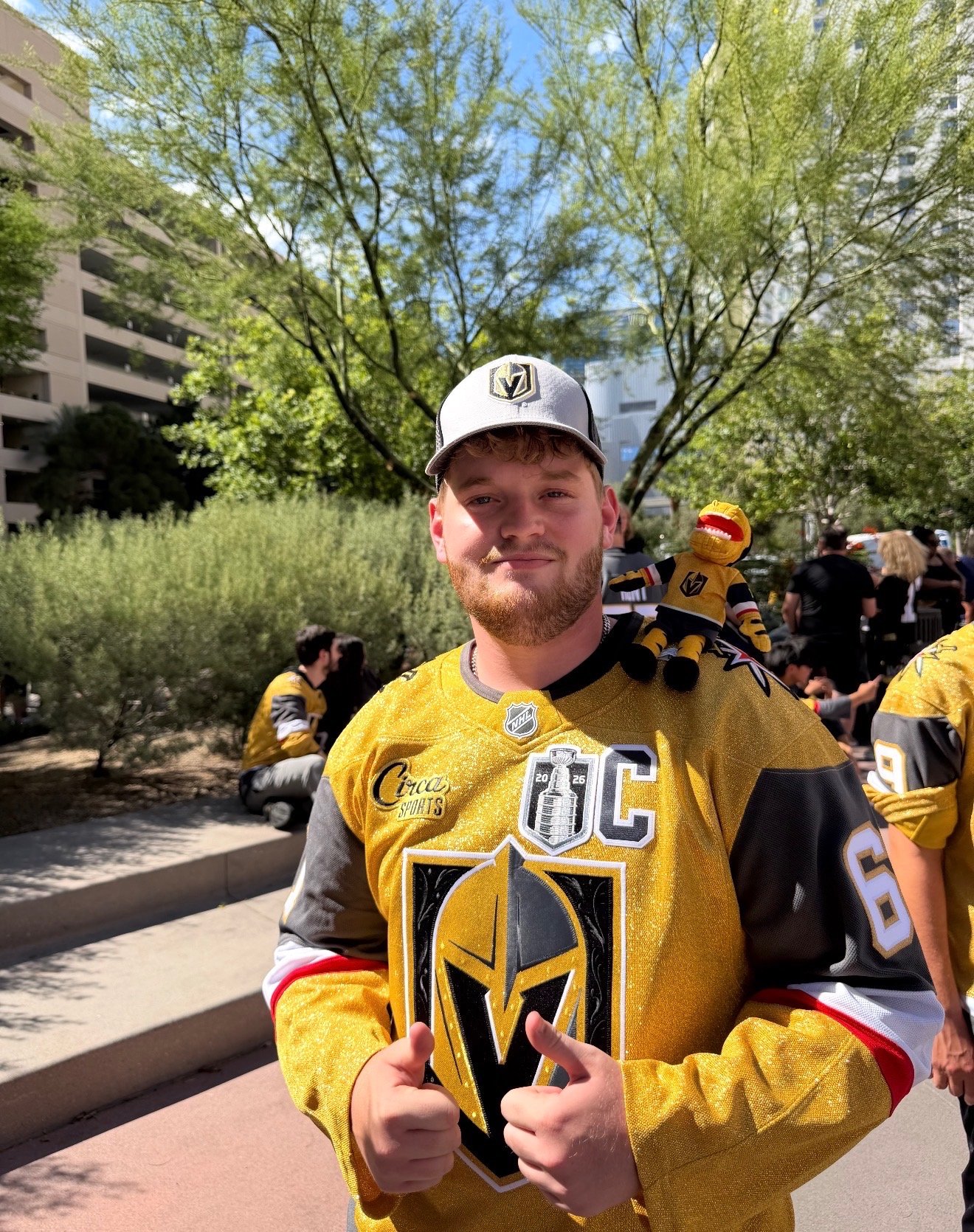A smiling hockey fan wearing a gold Vegas Golden Knights jersey and cap gives two thumbs up outdoors, with a small team mascot plush perched on his shoulder.
