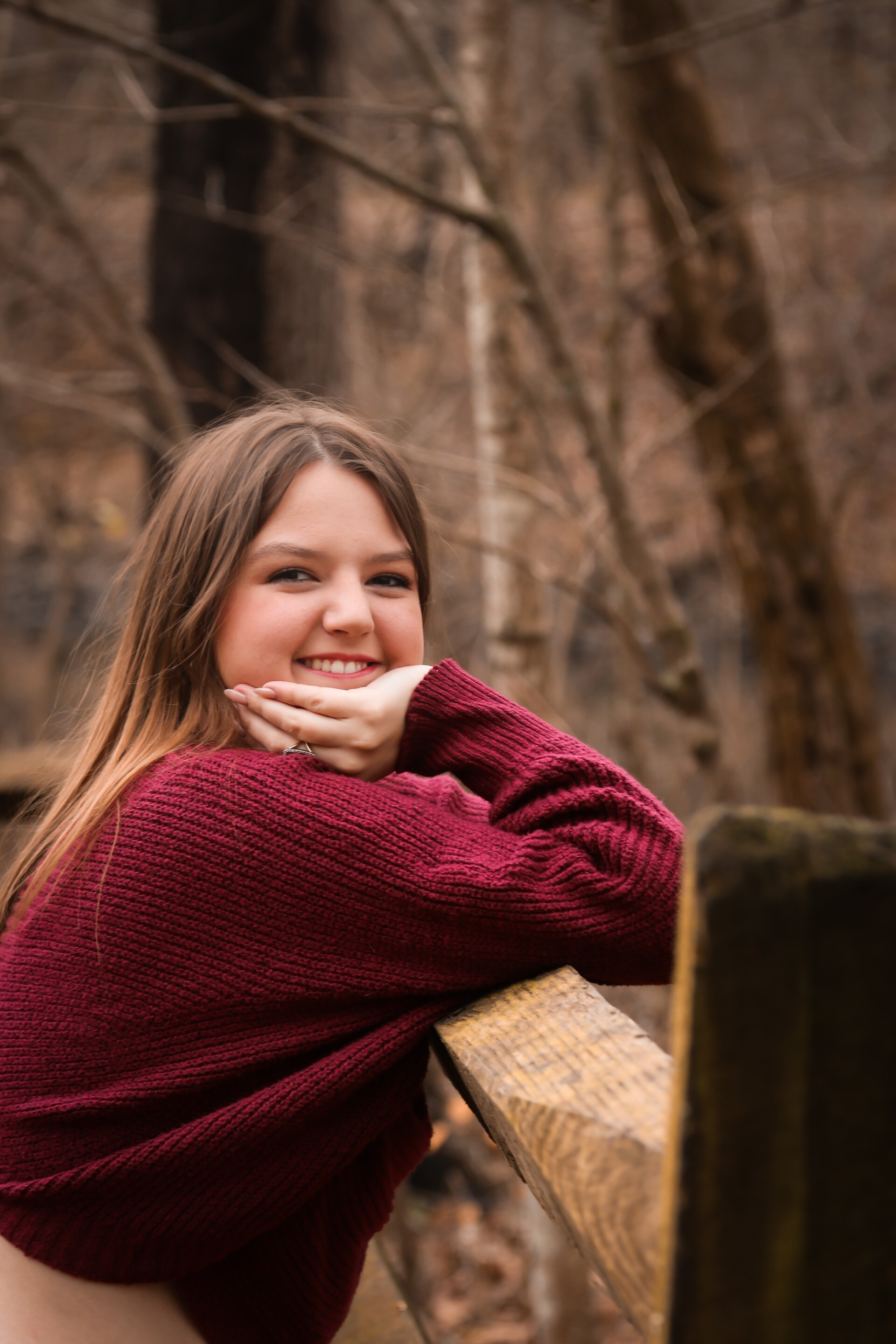Young woman in a burgundy knit sweater smiling while leaning on a wooden railing in a wooded outdoor setting. She rests her chin on her hands, with bare trees and a softly blurred natural background creating a warm, peaceful portrait.