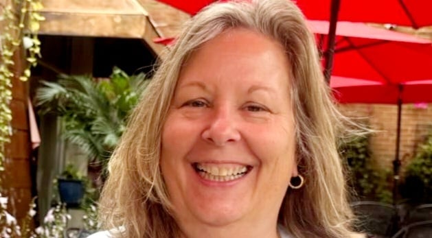 Close-up portrait of a smiling woman with shoulder-length blonde hair and hoop earrings, seated outdoors with greenery and red patio umbrellas in the background.