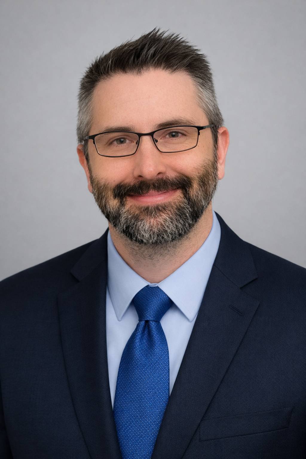 Professional headshot of a smiling man with short salt-and-pepper hair, a neatly trimmed beard, and glasses. He is wearing a navy suit, light blue shirt, and blue tie, posed against a light gray background with a friendly, approachable expression.