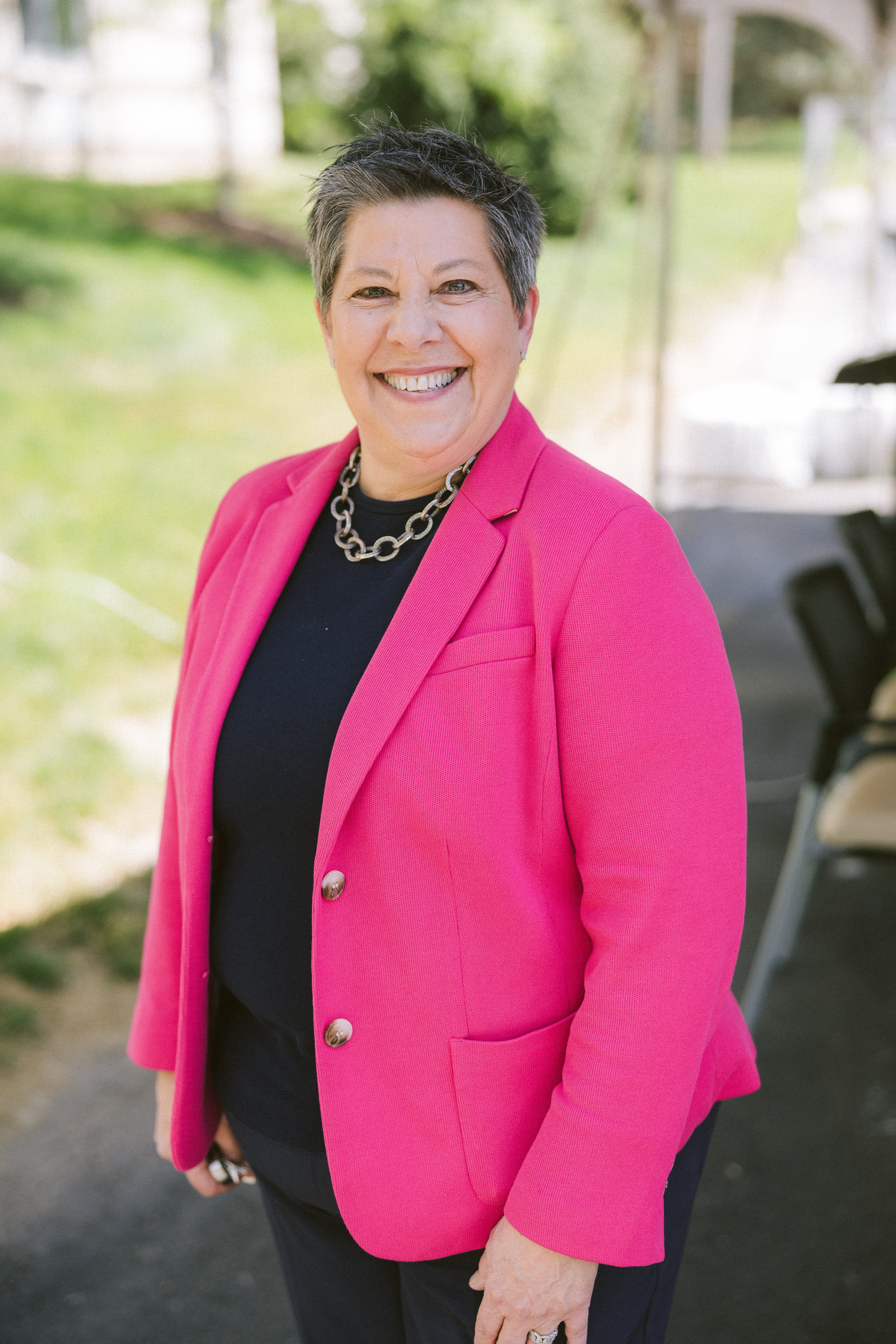 Smiling professional wearing a bright pink blazer over a dark top, standing outdoors with a softly blurred background. She has short salt-and-pepper hair, a silver chain necklace, and a warm, approachable expression, creating a polished and welcoming portrait.