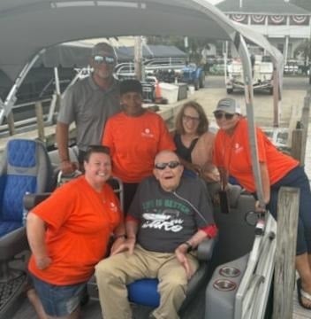 Older man wearing sunglasses smiles from a seat on a pontoon boat, surrounded by five companions gathered around him at a marina.